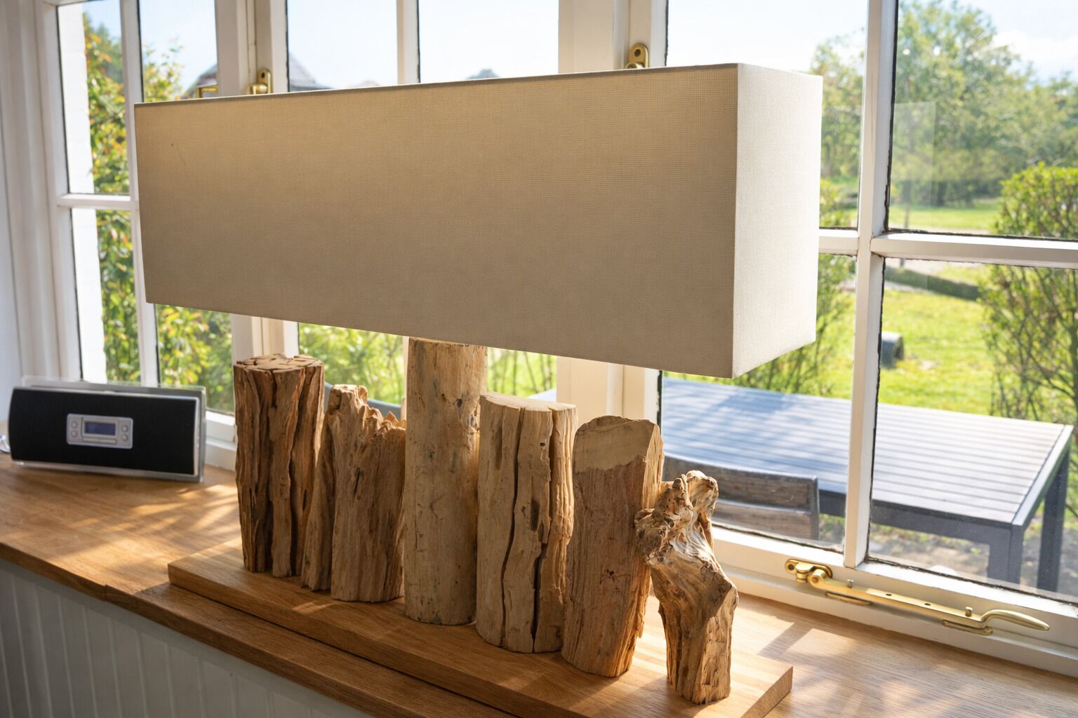 A modern lamp with a wooden base and a white lampshade on a wooden surface near a window.