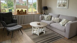 A cozy living room with a grey sofa, a grey armchair, a round coffee table, and a window with a view of greenery and buildings. - Bright apartment with direct access to the garden (Sankt Peter-Ording)