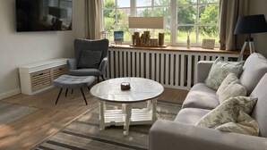 A living room with a grey armchair, a round coffee table, a sofa with floral cushions, a TV mounted on the wall, and a window with curtains. - Bright apartment with direct access to the garden (Sankt Peter-Ording)