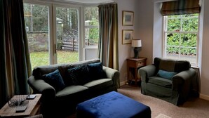 Una sala de estar con sofá, sillón y reposapiés, ventanas amplias con cortinas, y una lámpara colgante.