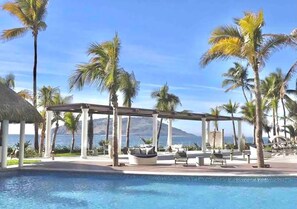 A poolside area with a pergola, lounge chairs, and palm trees. - Condos CAM (Mazatlán)