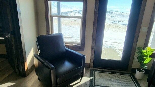 A room with a black leather chair, a view of a snowy landscape through a window, and a potted plant near the window. - Beautiful brand new house with breathtaking Shining Mountain views. (Ennis)