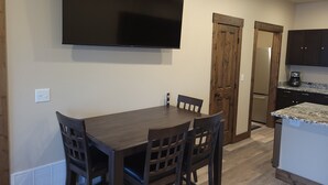 A dining area with a wooden table and chairs, a flat-screen TV mounted on the wall, and a kitchen area with dark cabinets and a countertop. - Beautiful brand new house with breathtaking Shining Mountain views. (Ennis)