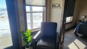 A room with a large window, a black leather chair, a potted plant, and a bed with a blue blanket. - Beautiful brand new house with breathtaking Shining Mountain views. (Ennis)