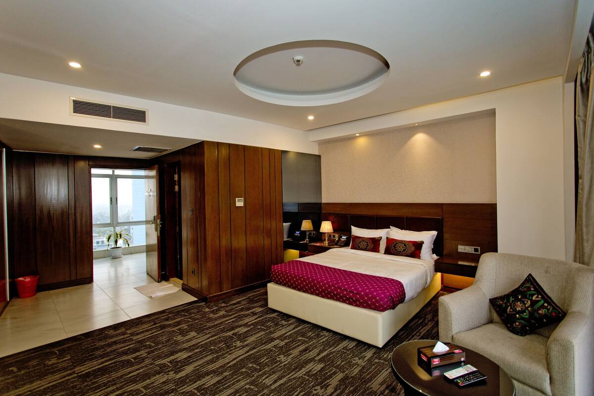 A hotel room with a bed, a chair, a nightstand, a lamp, and a view of the outdoors.