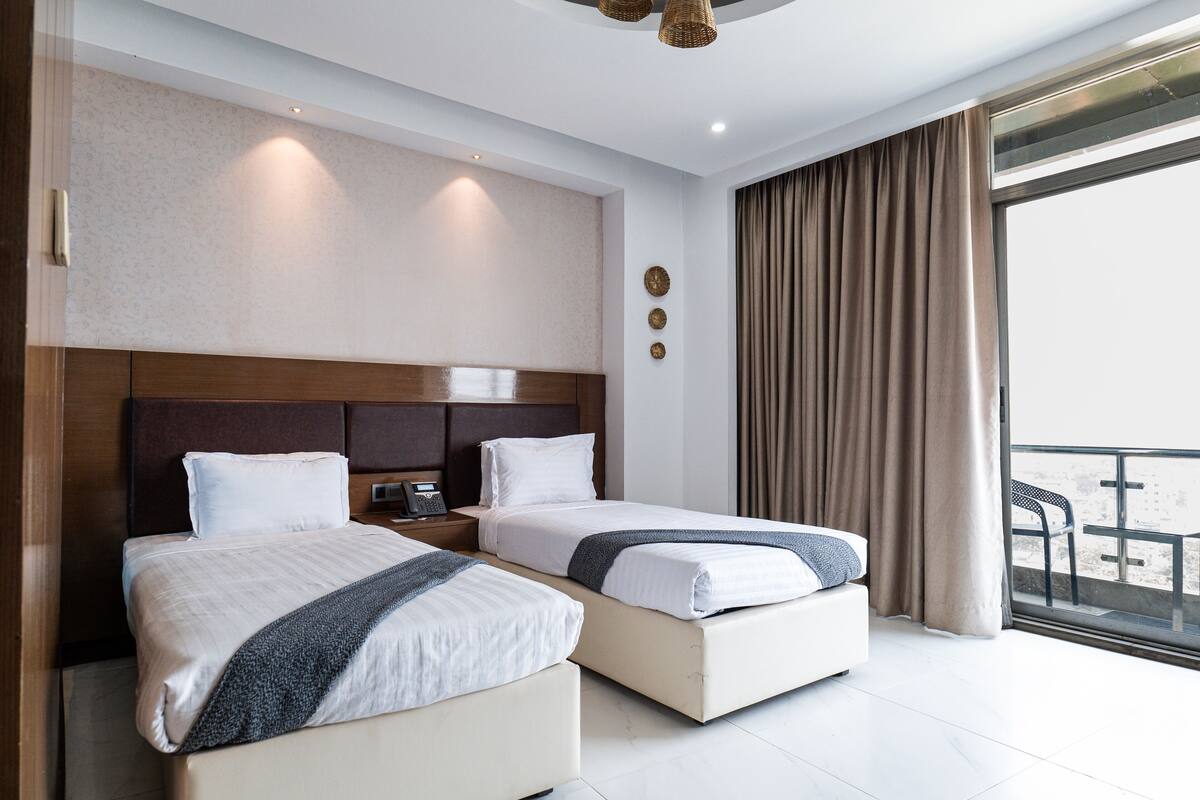 A hotel room with two beds, a large window with curtains, a wooden headboard, and a small table with a phone.