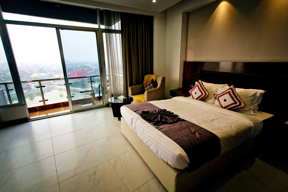 A hotel room with a large bed, a chair, a nightstand, and a view of the city through large windows.