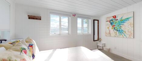 A bright, white room with a large bed, a small table, and a window with a view of the sea.