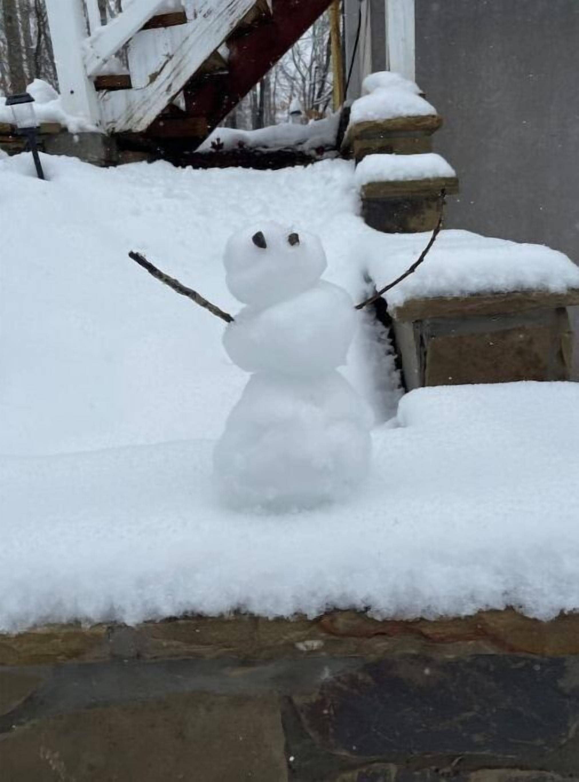 Un bonhomme de neige, coiffé d’un chapeau fait d’un bâton et doté de bras également faits de bâtons, se tient sur une corniche de pierre recouverte de neige.