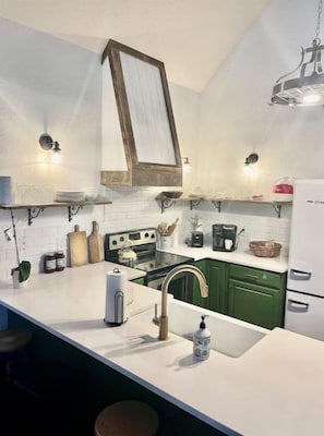 Una cocina con encimeras blancas, armarios verdes, una lámpara colgante y un refrigerador blanco.
