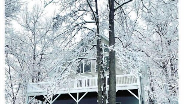 Una casa de dos pisos con un porche, rodeada de árboles cubiertos de nieve.
