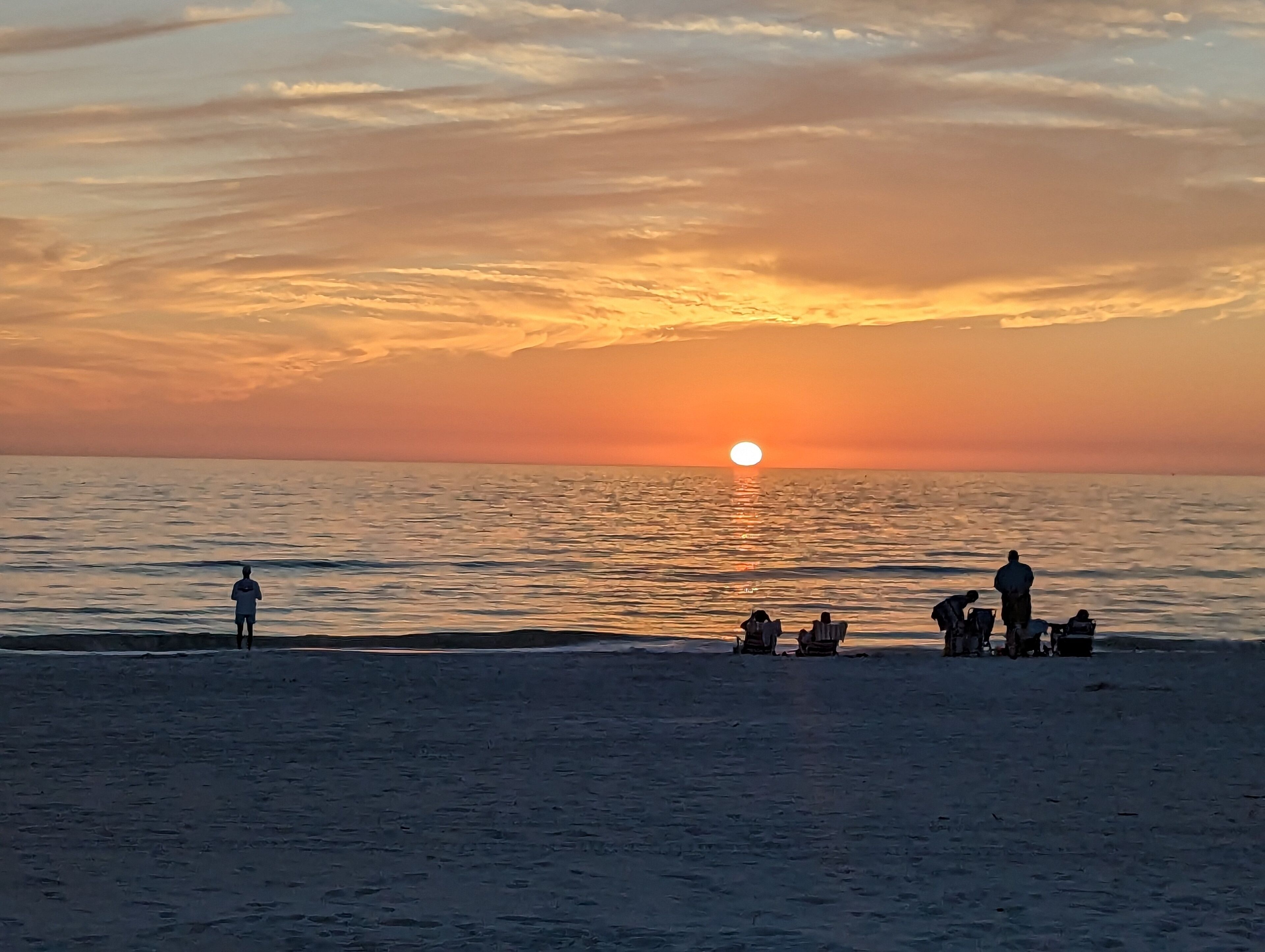 A beach scene with a sunset over the ocean, people standing and sitting on the sand, and a clear sky.