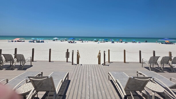 Een houten terras met lounge stoelen en uitzicht op een strand met parasols en mensen.