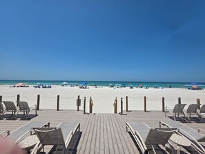 Una terraza de madera con sillones de descanso y vista a una playa con sombrillas y gente.
