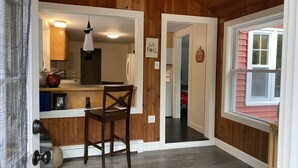 Una habitación con paredes de madera, una mesa de comedor de madera con sillas, una zona de cocina con un refrigerador y una ventana que ofrece vistas a un edificio rojo.