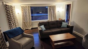 Sala de estar con sofá, sillón y mesa de centro. Hay una ventana con cortinas, piso de madera y vista al exterior.