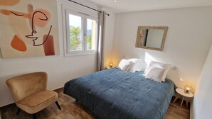 Un dormitorio con cama, una silla, un ventanal y un espejo adosado a la pared.