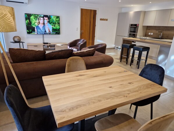 A modern living room with a wooden dining table, brown sofa, and a TV mounted on the wall.