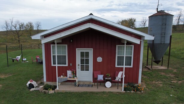 Une cabane en bois rouge, avec une porte et des fenêtres blanches, une véranda avec deux chaises, et un réservoir d’eau en arrière-plan.