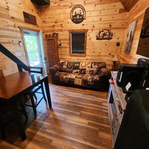 A cabin interior with a wooden floor, a sofa with a horse-themed blanket, a dining table, and a kitchen area with a coffee maker. - Windstorm Cabin (Marshall)