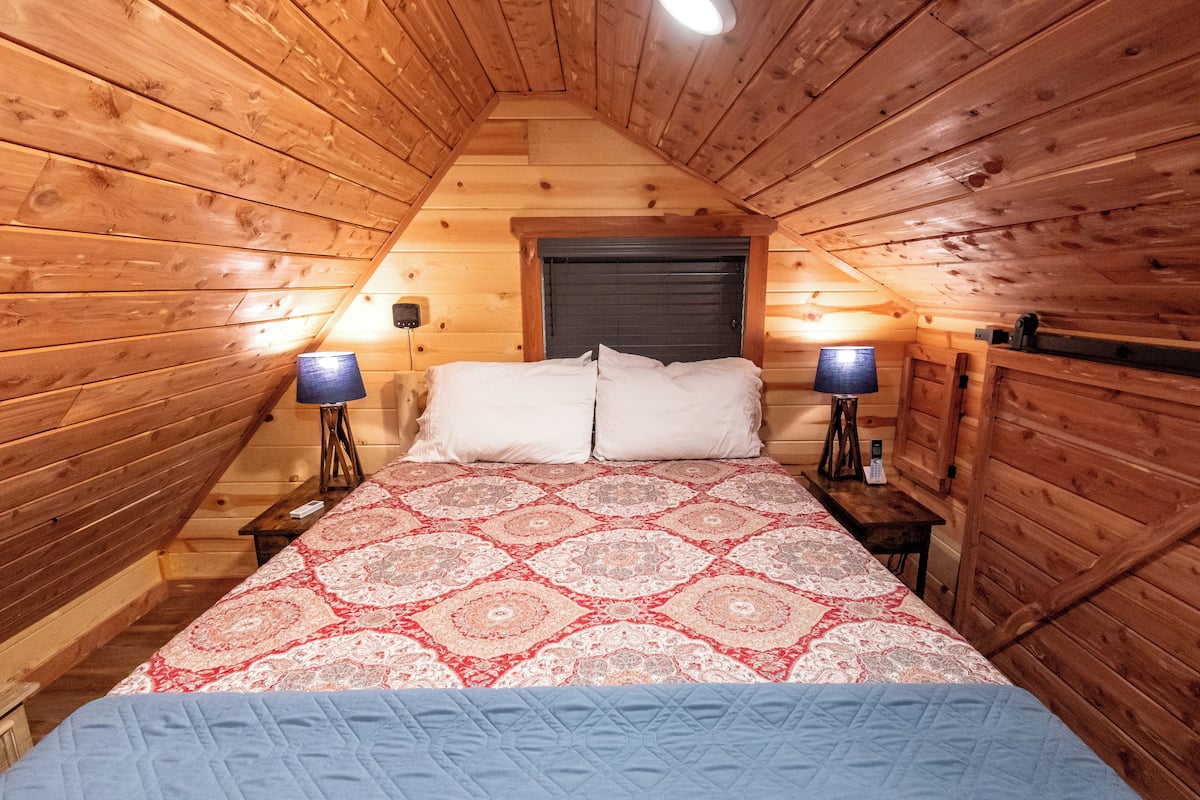 Une chambre confortable située sous les toits, avec un plafond en pente en bois, un grand lit recouvert d’une literie à motifs, deux lampes de chevet et une tête de lit sombre.