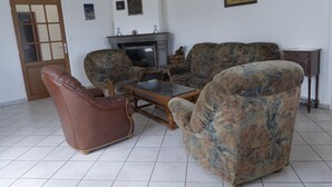 Olohuone, jossa on takka, kaksi kuviollista sohvaa, ruskea nahkainen nojatuoli ja pieni puinen kaappi.