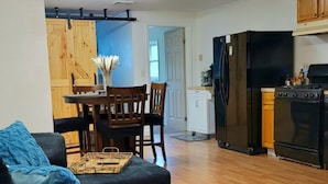 A living room with a dark sofa, a dining area with a wooden table and chairs, a kitchen with a refrigerator and stove, and a ceiling fan.