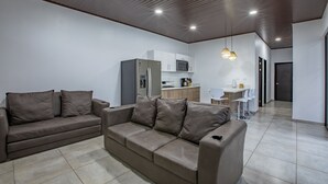 A modern living room with a sofa, a dining area with chairs, and a kitchen in the background.