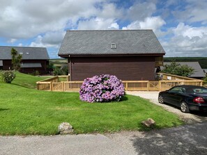 A wooden cabin with a deck, surrounded by a well-maintained lawn and a large bush of purple flowers.