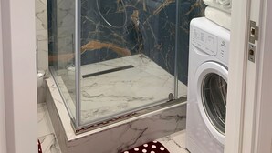 Une salle de bain moderne dotée d’une douche à parois de verre, de murs en marbre et d’un lave-linge encastré.
