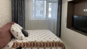 Une chambre à coucher avec un lit, un téléviseur fixé au mur, un lustre et une fenêtre avec des rideaux.