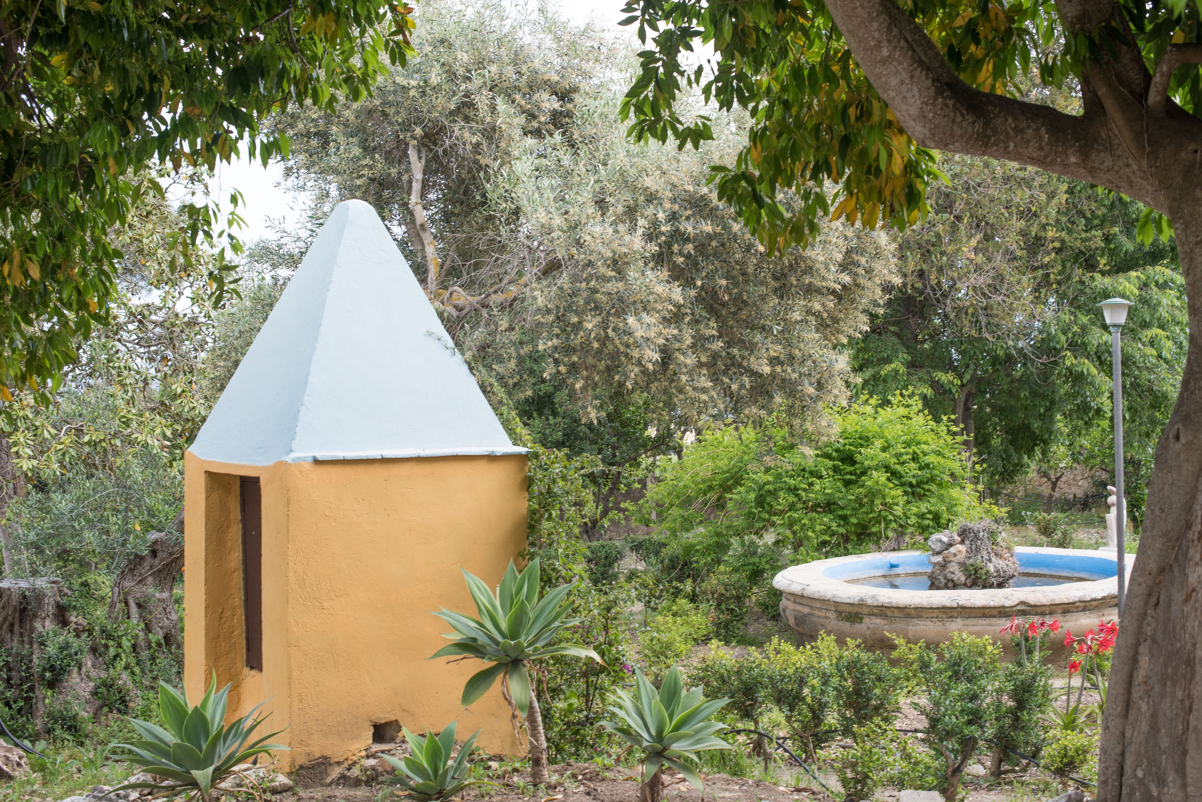 Un petit bâtiment jaune, surmonté d’un toit conique, entouré d’une végétation luxuriante et agrémenté d’une petite fontaine.