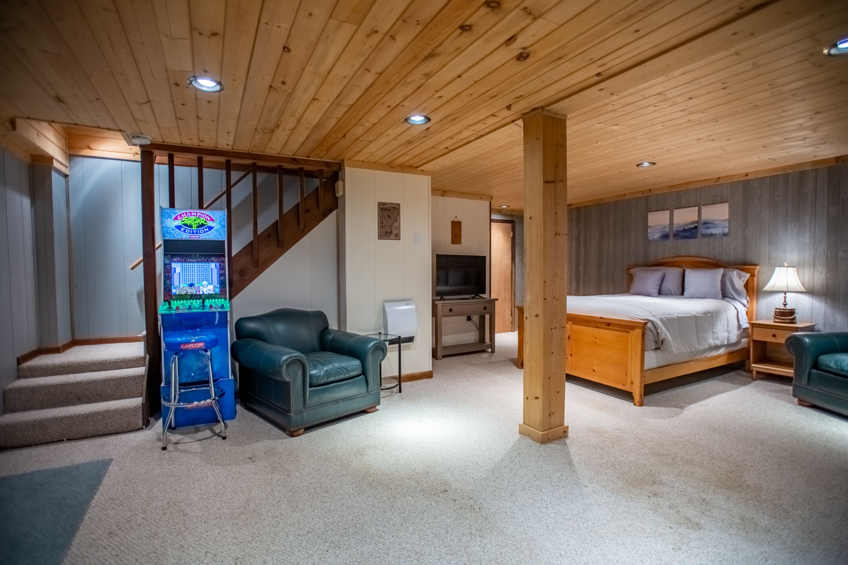 Quarto com cama de madeira, poltrona verde, escrivaninha pequena e um videogame.