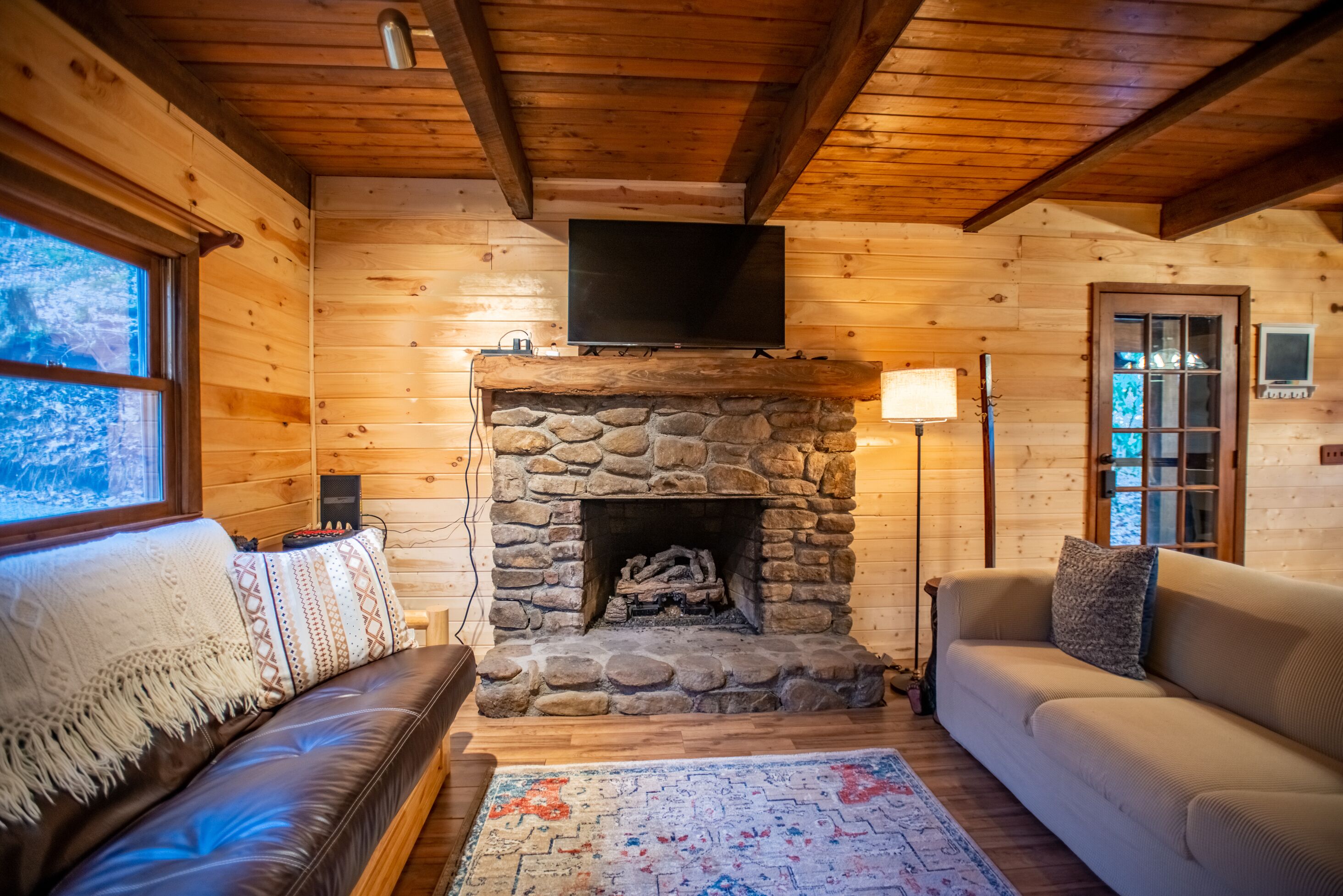 A cozy cabin interior with a stone fireplace, leather sofas, and a flat-screen TV mounted above the fireplace.