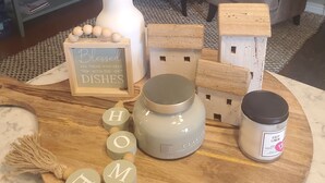 Un coin cuisine agrémenté d’une plante décorative, d’une planche à découper en bois avec des bougies, d’un panneau et d’une bougie parfumée dans un écrin en lin.