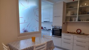 A modern kitchen with white cabinets, a dining table with a patterned tablecloth, and a window with frosted glass. - Comfortable apartment just minutes from the city (Su Planu)