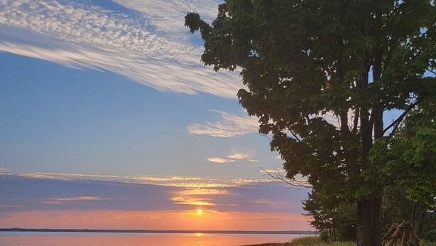 Een camping met een ronde vuurkuil en picknicktafels, omringd door bomen en uitzicht op de zonsondergang over het water.