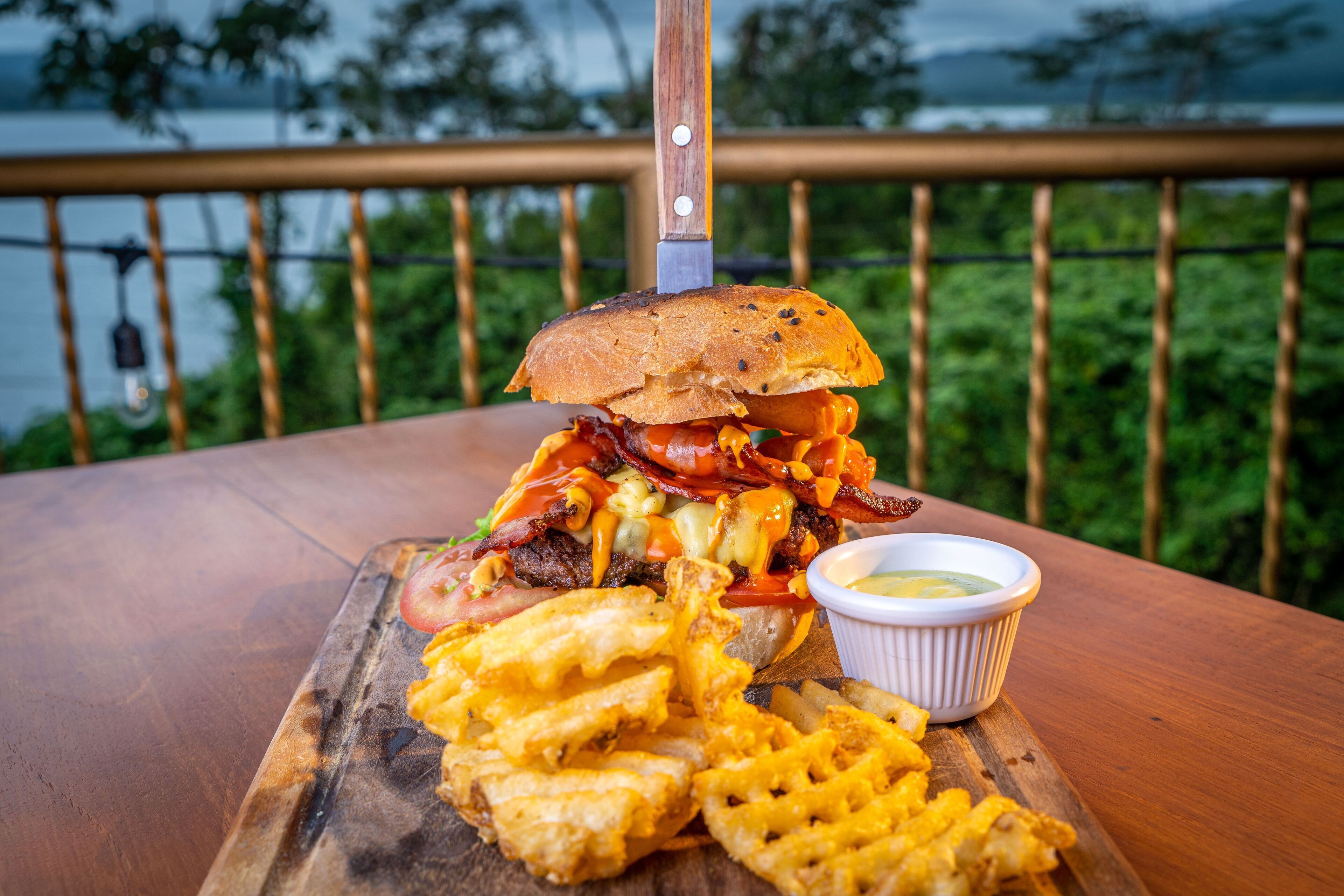 A burger with bacon, cheese, and tomato on a wooden board, accompanied by fries and a dipping sauce, set on a table with a railing and greenery in the background.