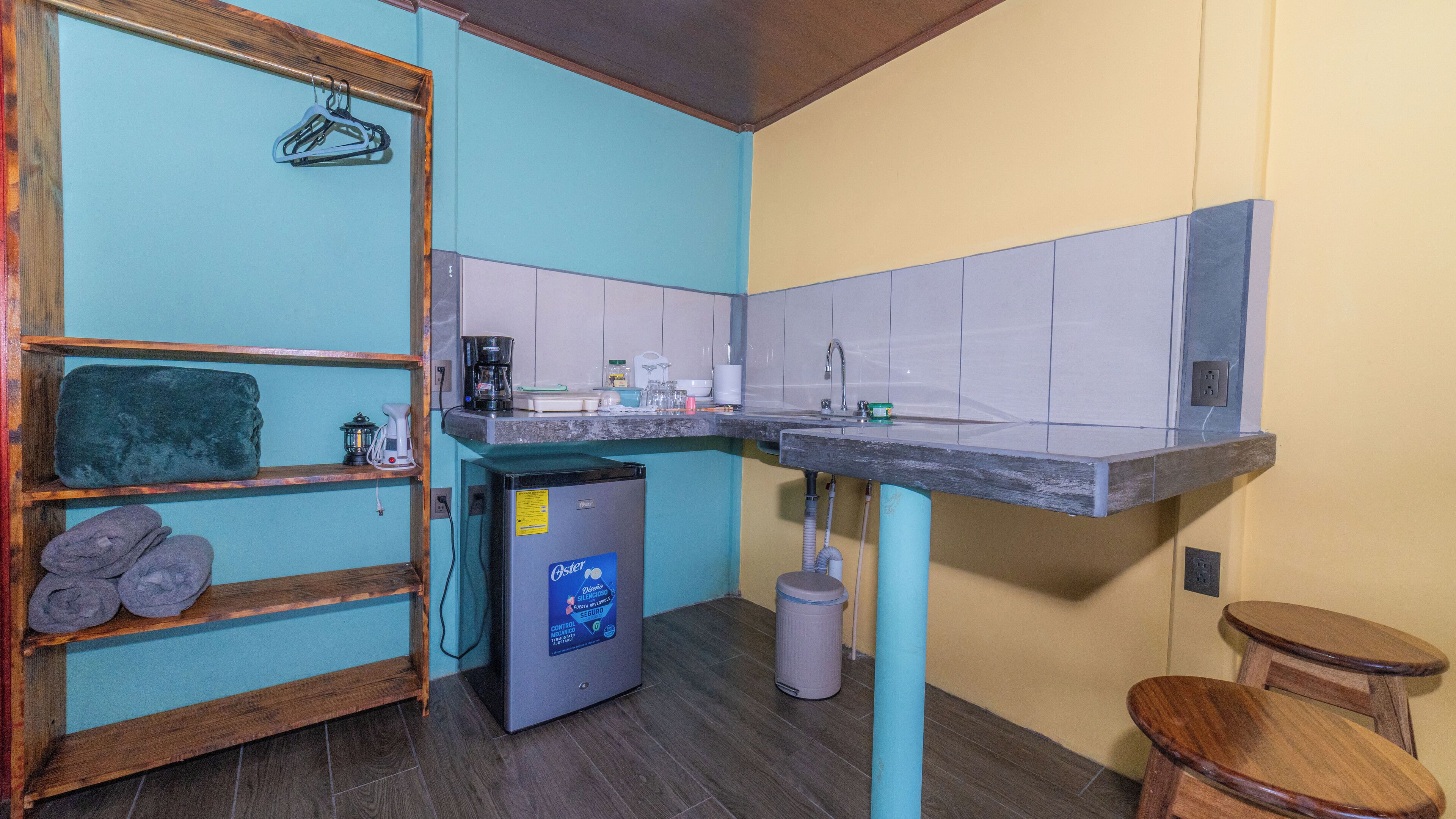 A compact kitchen with a sink, a coffee maker, and a small refrigerator. There are wooden shelves with towels and a hanging clothes rack.