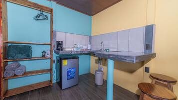 A compact kitchen with a sink, a coffee maker, and a small refrigerator. There are wooden shelves with towels and a hanging clothes rack.