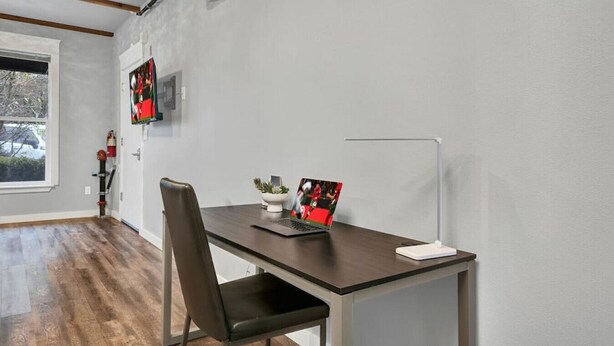 A modern office space with a desk, chair, and a laptop. There is a window with a view of trees and a fire extinguisher on the wall. - Extended Stay Comfort Fast Wi-Fi  Downtown! (Tacoma)