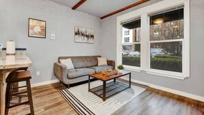 Un salon avec un canapé gris, une table basse, une fenêtre donnant sur l’extérieur et une petite table d’appoint sur laquelle est posée une bougie.