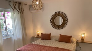 Ein Schlafzimmer mit einem Bett, zwei Nachttischen mit Lampen, einem runden Spiegel, einem Fenster mit Vorhängen und einer Wandklimaanlage.