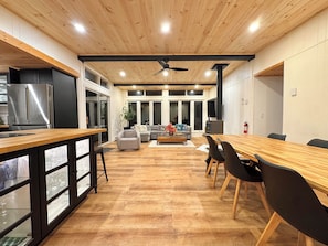 Une cuisine moderne avec un plafond en bois, un coin repas avec une longue table et des chaises, et un salon avec un canapé et une table basse.