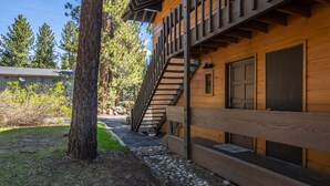 A wooden cabin with a staircase leading to a door, surrounded by trees and a gravel path. - Cozy Cabin-Style Condo in Central Location (Mammoth Lakes)