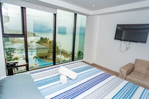 Une chambre d’hôtel moderne dotée d’une grande fenêtre donnant sur une piscine et l’océan, d’un téléviseur à écran plat fixé au mur et d’un lit agrémenté d’un tapis rayé.