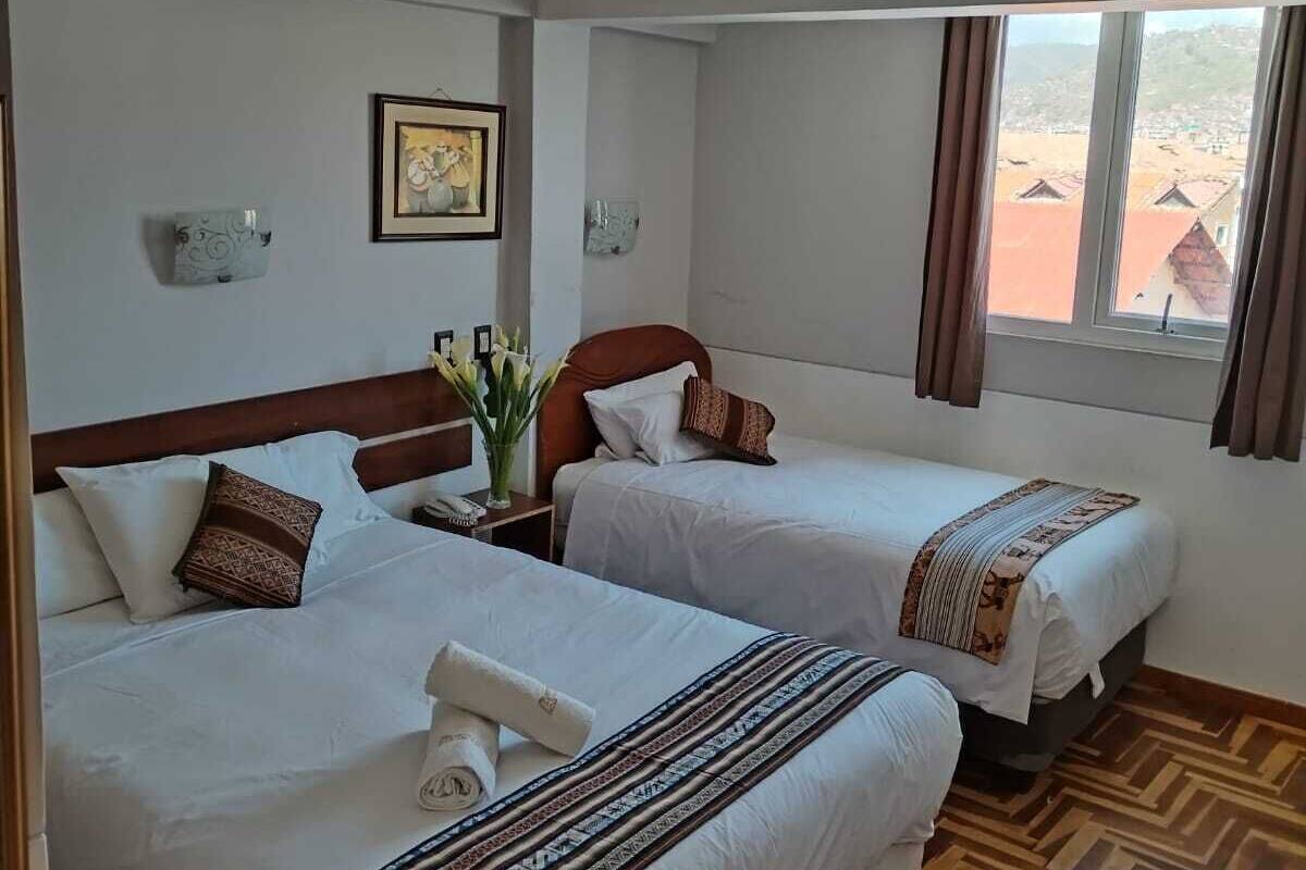 A hotel room with two beds, a wooden floor, a window with curtains, a framed picture on the wall, and a small table with a vase of flowers.