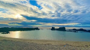 Uma praia com águas calmas, formações rochosas ao longe e céu claro.