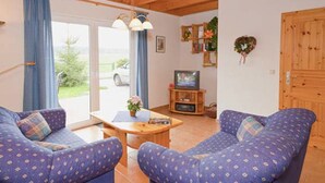 Un salon chaleureux avec deux canapés bleus à motifs, une table basse en bois, un téléviseur posé sur un meuble en bois, et une vue sur une voiture garée à l’extérieur, visible à travers une porte vitrée.