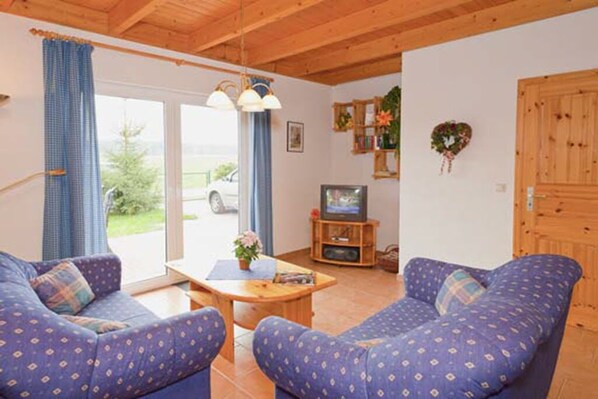 A cozy living room with two blue patterned sofas, a wooden coffee table, a television on a wooden stand, and a view of a car outside through a glass door. - Vacation home in a quiet location, 200m from the nature reserve lake, Mönchshof 13b, with WLAN (Eldetal)
